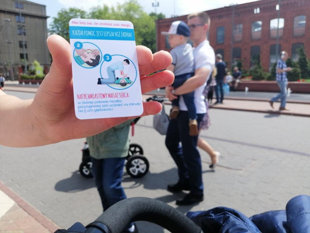 Zdjęcie przechodniów w Manufakturze. Na pierwszym planie dłóń z ulotką.